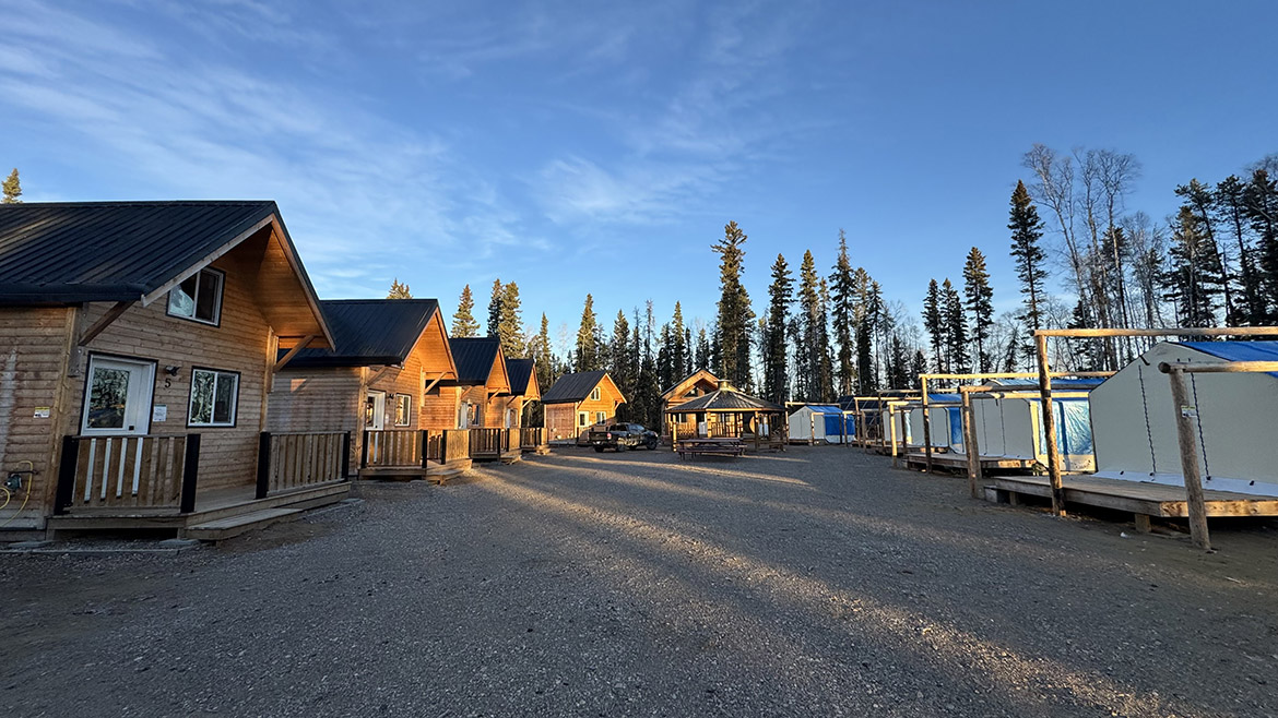 A wide shot of five small cabins, one big cabins, a gazebo, and five tents around a clearing in the forest.