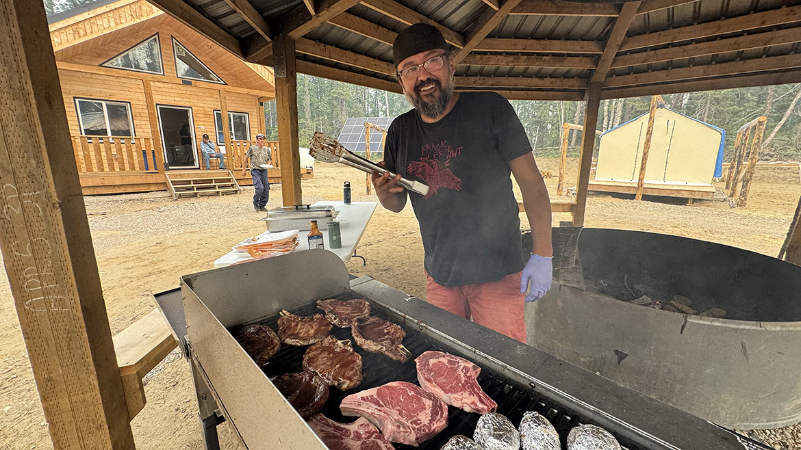 A bearded man smiles while barbecuing under a gazebo. A tent and large cabin are seen in the background.